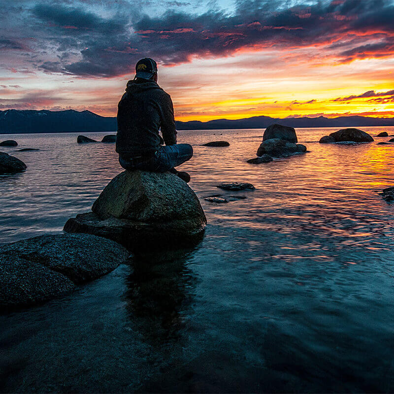 Photo of a man sitting by himself near a lake at sunset